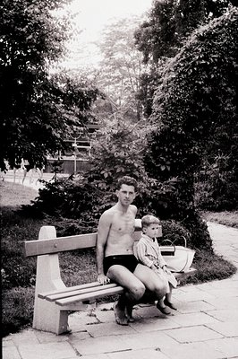 A man, bare-chested and wearing swim trunks, sits on a concrete bench with a young child dressed in a light-colored outfit. L...