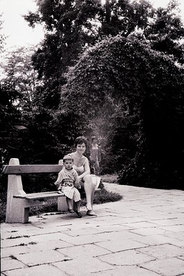 A mother and child sit on a stone bench, framed by dense foliage. The child wears a short patterned dress, and the woman is i...