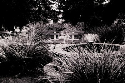 A woman and child seated on a bench overlook a circular fountain within a manicured garden. The architecture suggests a Europ...