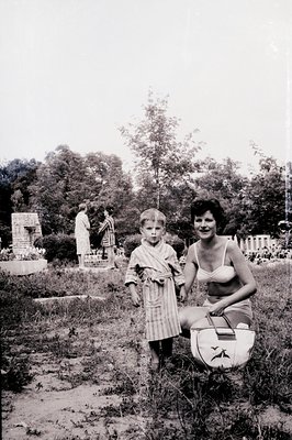 A young boy in a striped robe stands with a woman in a vintage bikini, seated near a travel bag. A classical ruin/statue is v...