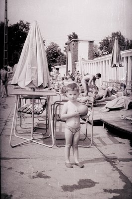 A young boy in swimming trunks stands holding a ball near a poolside patio set. A wooden deck extends into a swimming pool ar...