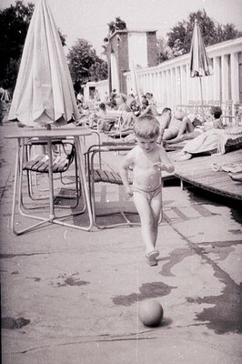 A young boy, scantily clad, runs on a wet concrete patio toward the camera, a ball at his feet. Behind him, a poolside scene ...
