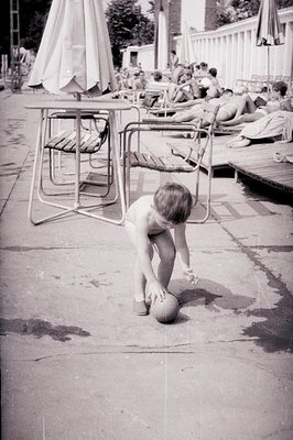 A young boy, partially obscured by patio furniture, crouches playfully near a ball on a concrete terrace. Behind him, figures...