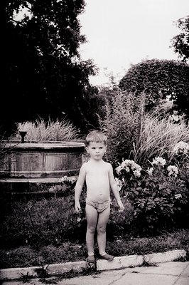 A young boy stands barefoot, partially obscured by foliage, in a formal garden setting. He wears only briefs. A woman's back ...