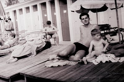 A candid poolside scene: A man and young boy relax on a wooden deck, while a young woman sunbathes nearby. Architectural deta...