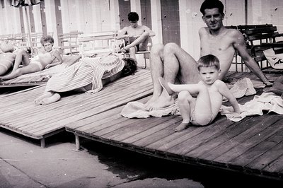 A vintage monochrome photo captures a family scene on a wooden boardwalk. A man sits with a young boy, both nude; a woman lie...