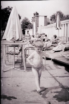 A wet, young boy in patterned swimming trunks poses playfully on a concrete poolside. Several sunbathers relax on deck chairs...