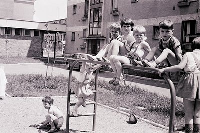 Group of children play on a seesaw in a concrete courtyard, likely a Soviet-era housing complex. The architecture suggests Ea...