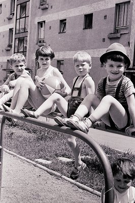 Four boys playfully perch on a metal playground structure in front of a block of apartments. Distinctive clothing – overalls,...