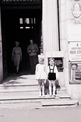 Two young girls, one in a dress & bow, the other in pinafore & suspenders, stand on stone steps. Adults exit a building behin...