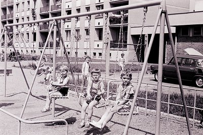 Three boys seated on swings in a public playground. Housing block architecture visible in the background, indicative of post-...