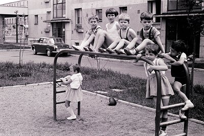Group of boys perched on a gymnasium structure, a girl below. Mid-century apartment complex backdrop; a dark sedan parked nea...