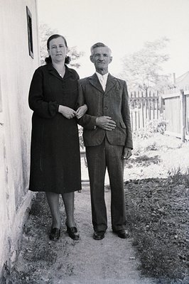 A formally dressed elderly couple stands outdoors, leaning against a weathered wall. The woman wears a dark, knee-length dres...