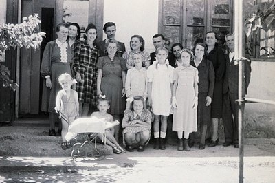 A formal group portrait; ten individuals and three children pose outside a building with a covered entryway. Clothing suggest...