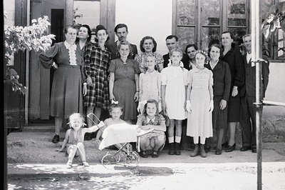 A large family group poses outdoors, likely in front of a home or institutional building. Women in mid-century dresses and me...