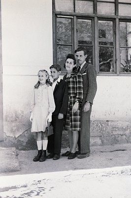 A family portrait: a girl, boy, and parents stand before a building with visible architectural details. The girl wears a dres...