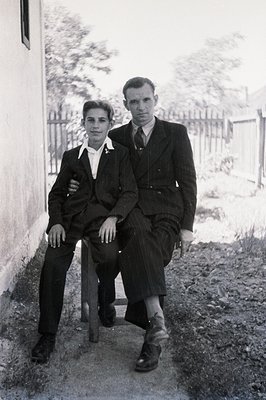 Formal portrait of a man and a young man seated outside a building, likely a home. The men wear dark suits, ties, and polishe...