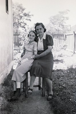 Two women sit outdoors, posed near a plain, stucco wall. Both wear modest, 1940s-style dresses and sturdy shoes. One woman’s ...