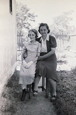 A young woman in a dress and older woman in a collared shirt sit side-by-side on simple stools. The setting appears to be a c...