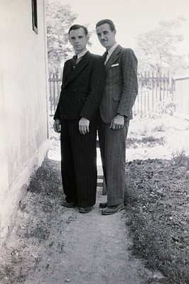 Two men stand beside a stone wall, formally attired in 1940s-style suits. The man on the left wears a dark double-breasted su...