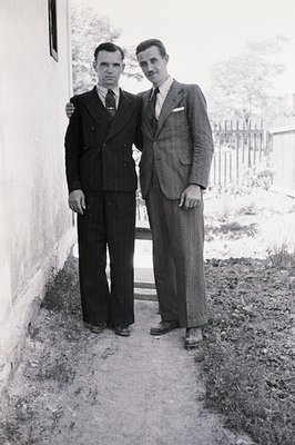 Two men in well-tailored suits stand closely together near a weathered stone wall and path. Formal attire suggests a special ...