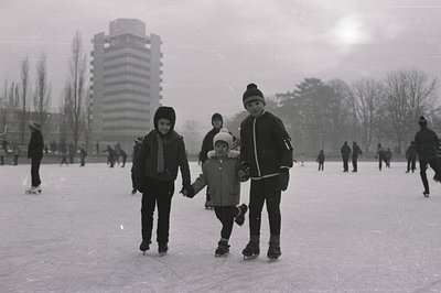 Three children ice skating on a frozen pond or lake, linked hands. A tall, brutalist apartment building rises in the backgrou...
