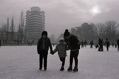 Monochrome image showing a man, presumably a father, supporting a young child learning to ice skate. A second child stands ne...