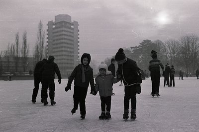 A group of children ice skating on a frozen pond, seemingly outdoors. A taller figure appears to be assisting the younger ska...