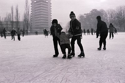 A young boy, assisted by two older children, learns to ice skate on a frozen pond. Background shows other skaters and a tall,...