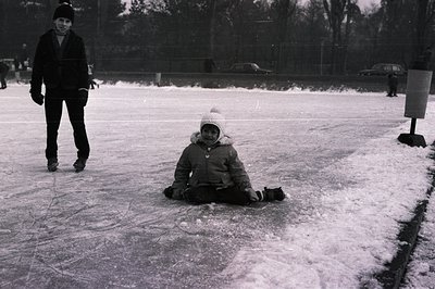Black & white photo showing a child seated on a snowy outdoor ice rink. A second individual is visible in the background, ska...
