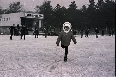 A young girl, bundled in a winter coat and hat, stands poised on skates on a frozen pond. Several other skaters populate the ...