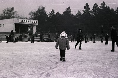 A monochrome image of an outdoor ice skating rink, likely in Eastern Europe. A child in a hooded coat stands center frame, se...