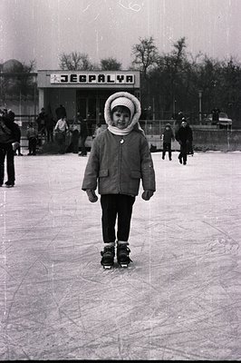 A young child, bundled in a warm coat & hat, stands confidently on an outdoor ice rink. "ZEBPALYA" signage visible in the bac...
