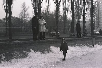 A monochrome street scene depicts a young boy in a dark coat walking along a snow-covered sidewalk. In the background, a coup...