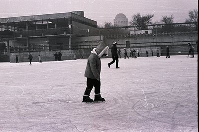 B&W image captures a public ice rink scene. Several figures skate and walk across the ice. A large, modern building with a cu...
