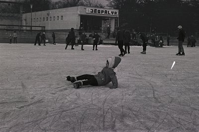 A child in winter clothing lies prone on a frozen outdoor ice rink, surrounded by other skaters. The "JEBPLYR" sign of a buil...
