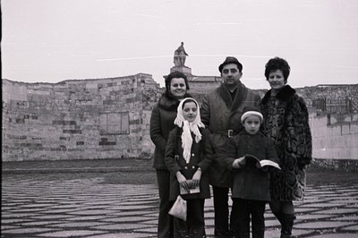 A family portrait from the 1960s or 70s. Four people stand outdoors before a stone wall topped with an ornate architectural s...