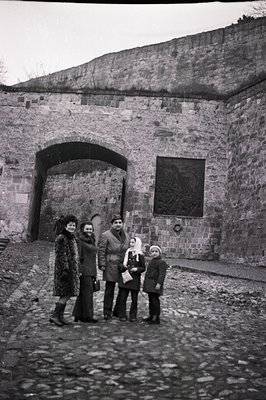 Family portrait before a stone archway, likely a fortress gate. Individuals are dressed in 1970s outerwear & bell-bottoms. A ...