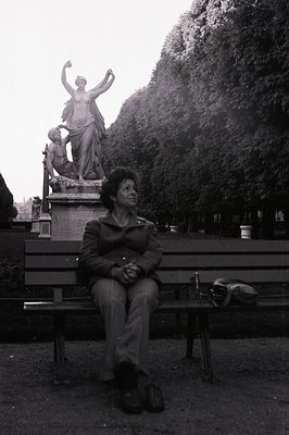 A seated individual wearing a dark jacket and corduroy trousers occupies a park bench before a monumental statue. Dense folia...