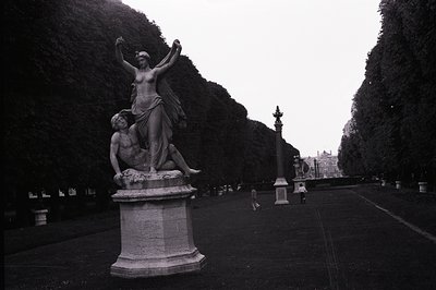 Ornate marble sculpture depicting a draped female figure supporting a fallen male figure. Formal, expansive park landscape wi...