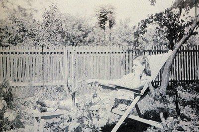 A young woman reclines in a wicker chair amidst overgrown foliage and a tall picket fence. The scene suggests a relaxed, priv...