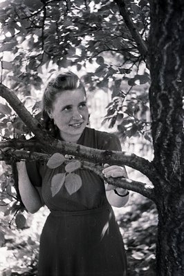 A young woman, seemingly in her late teens, smiles directly at the camera while leaning on a tree branch. She wears a modest,...
