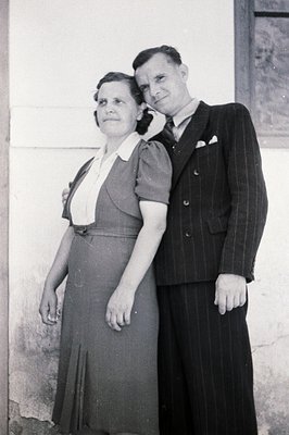A formally attired couple poses outside a stucco building. The woman wears a modest, knee-length dress with a contrasting col...