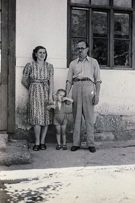 A formal portrait captures a family—a woman, man, and young child—standing outside a building. The man, wearing glasses, hold...