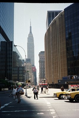 A view down a New York City street toward the Empire State Building, framed by distinctive mid-century architecture. A yellow...
