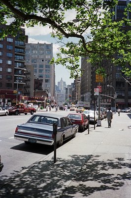 A 1970s-80s urban street scene in New York City. Classic cars line the curb; pedestrians populate the sidewalk and crosswalk....
