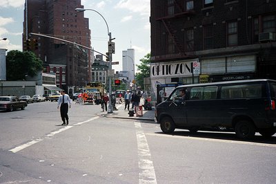 A bustling city street scene, likely NYC, circa 1970s or 80s. Construction work occupies a lane, with workers and equipment v...