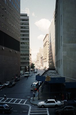 Looking down a city street framed by towering, modernist office buildings. Visible cars and pedestrian traffic suggest urban ...