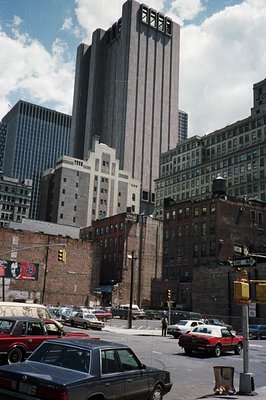 A view of a distinctive, tiered building in downtown New York City. Red brick buildings and a billboard flank the foreground....