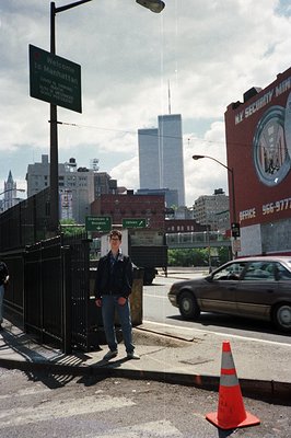 A man stands near a "Welcome to Manhattan" sign, overlooking a highway and the iconic World Trade Center towers. The photogra...